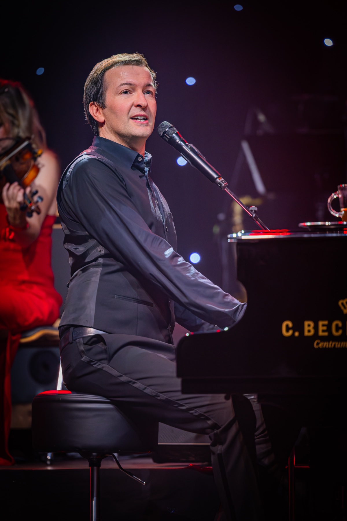 Alex Parker singt am Flügel, hinter ihm eine Geigerin im roten Kleid